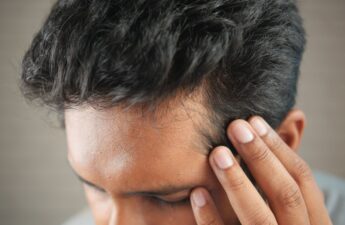 Man with dark hair holding his temple, showing hairline and early signs of hair loss near the forehead.